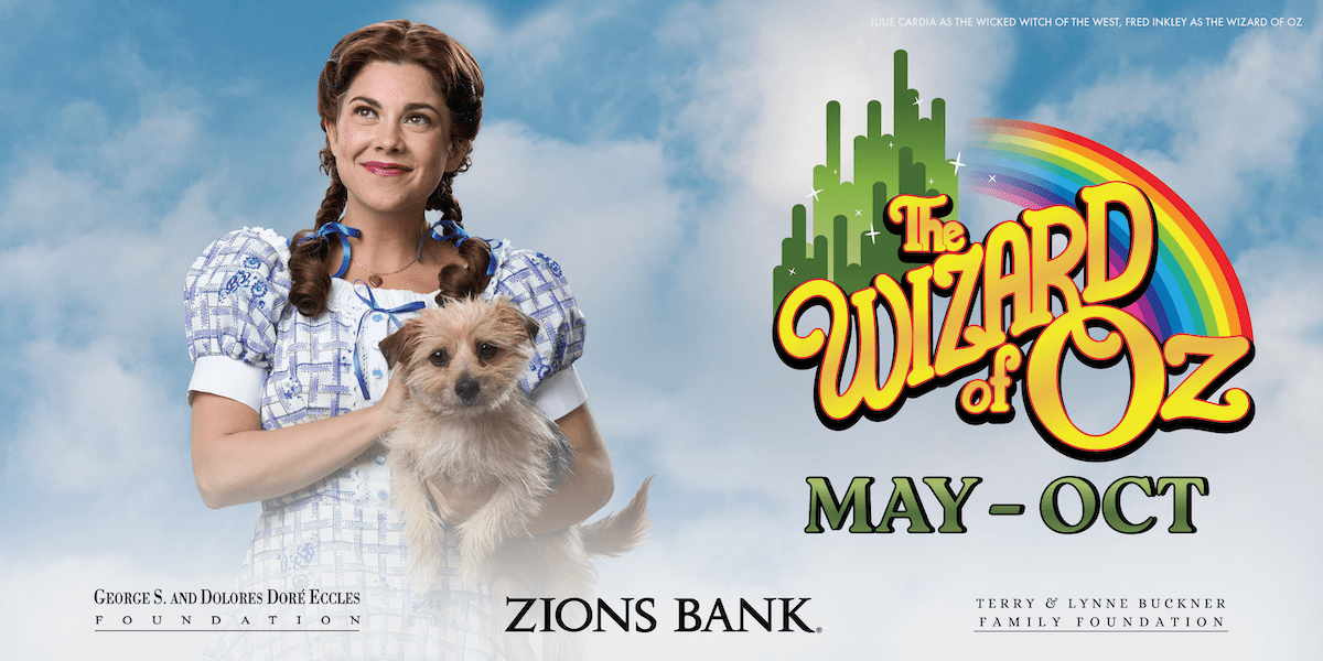 A promotional image for The Wizard of Oz at Tuacahn Amphitheatre. On the left, a young woman dressed as Dorothy smiles and holds a small tan dog, Toto, against a cloudy blue sky background. She wears a blue-and-white gingham dress with puffed sleeves and blue ribbons in her braided hair. On the right side, the show's title "The Wizard of Oz" appears in colorful text over a green silhouette of the Emerald City and a rainbow. Below the title, the run dates "May – Oct" are displayed. Sponsor logos for Zions Bank, the George S. and Dolores Doré Eccles Foundation, and the Terry & Lynne Buckner Family Foundation are also visible.