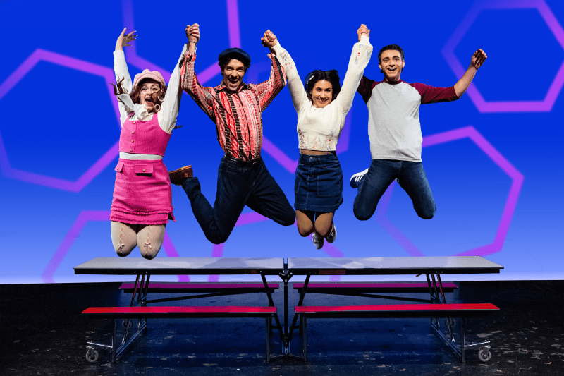 Image Description: Four young adult performers leap into the air with arms raised in excitement, standing atop a red and gray cafeteria-style lunch table. From left to right: the first actor wears a pink jumper dress over a white blouse with heart-shaped elbow patches and a pink beret; the second wears black pants, a red-and-white striped Western-style shirt, and a black hat; the third wears a white long-sleeved blouse with embroidered accents and a denim skirt; the fourth wears jeans and a gray T-shirt with maroon sleeves. They are mid-jump, smiling energetically against a bright blue background with large pink hexagon outlines, capturing a moment of youthful joy and camaraderie.