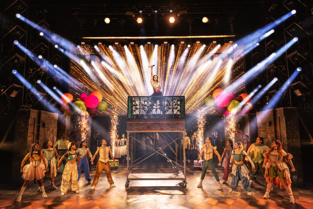 The cast of & Juliet performs a high-energy musical number on stage. Centered on a raised platform, a female performer (Juliet) stands triumphantly with one arm raised, holding a microphone. She is illuminated by dramatic golden pyrotechnics and surrounded by beams of white stage lights pointing outward. Below her, a diverse ensemble of actors stands in a V formation, gazing up at her with admiration and energy. The cast wears colorful, modernized Renaissance-inspired costumes. Large bunches of bright balloons flank the platform on either side. The stage background is filled with theatrical lighting trusses, creating a bold and celebratory concert atmosphere.
