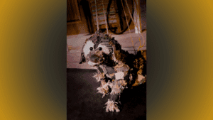 A handmade dog puppet sits on a dark floor, lit by theatrical lighting. The puppet is constructed from patchwork fabric in earthy tones—black, brown, beige, and rust—with visible seams, buttons, and frayed edges giving it a worn, homemade look. It has wide, round eyes, a button-like nose, and is controlled by ropes or strings from above, indicating it's a marionette. In the background, parts of a rustic stage set are visible, including wooden paneling and a wagon wheel, evoking an early 20th-century setting.
