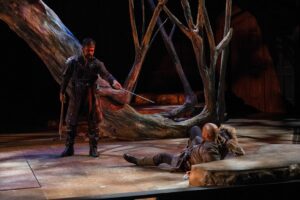 Walter Kmiec (left) as Macbeth and Lavour Addison as Macduff perform a climactic scene in the Utah Shakespeare Festival’s 2025 production of Macbeth. Onstage, Macbeth stands in dark armor and a long tartan robe, sword extended toward a fallen Macduff, who lies on the ground looking up at his opponent. The set features a massive, twisted tree trunk arching dramatically behind them, creating a stark, eerie forest aesthetic. Lighting casts moody shadows across the floor, enhancing the tension of the confrontation.