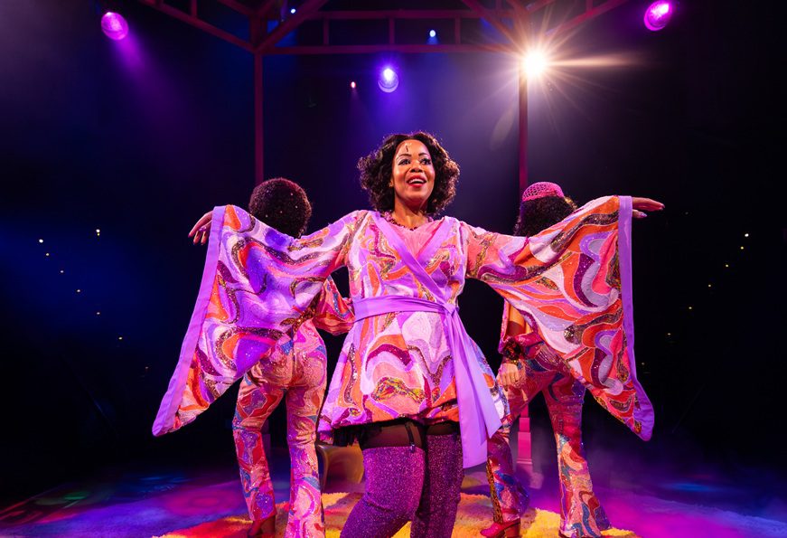 The image depicts a scene from a Hale Centre Theatre production of "Sister Act." At the center, a performer is joyfully extending her arms while wearing a vibrant, swirling-patterned outfit. Her costume features wide sleeves and a light purple belt. She stands confidently on a colorful, lit stage with two other performers in similar colorful attire visible in the background. The stage is lit with purple and pink spotlights creating a lively and dynamic atmosphere. Tiny aisle lights can be seen in the background, indicative of the theatre in the round setting.