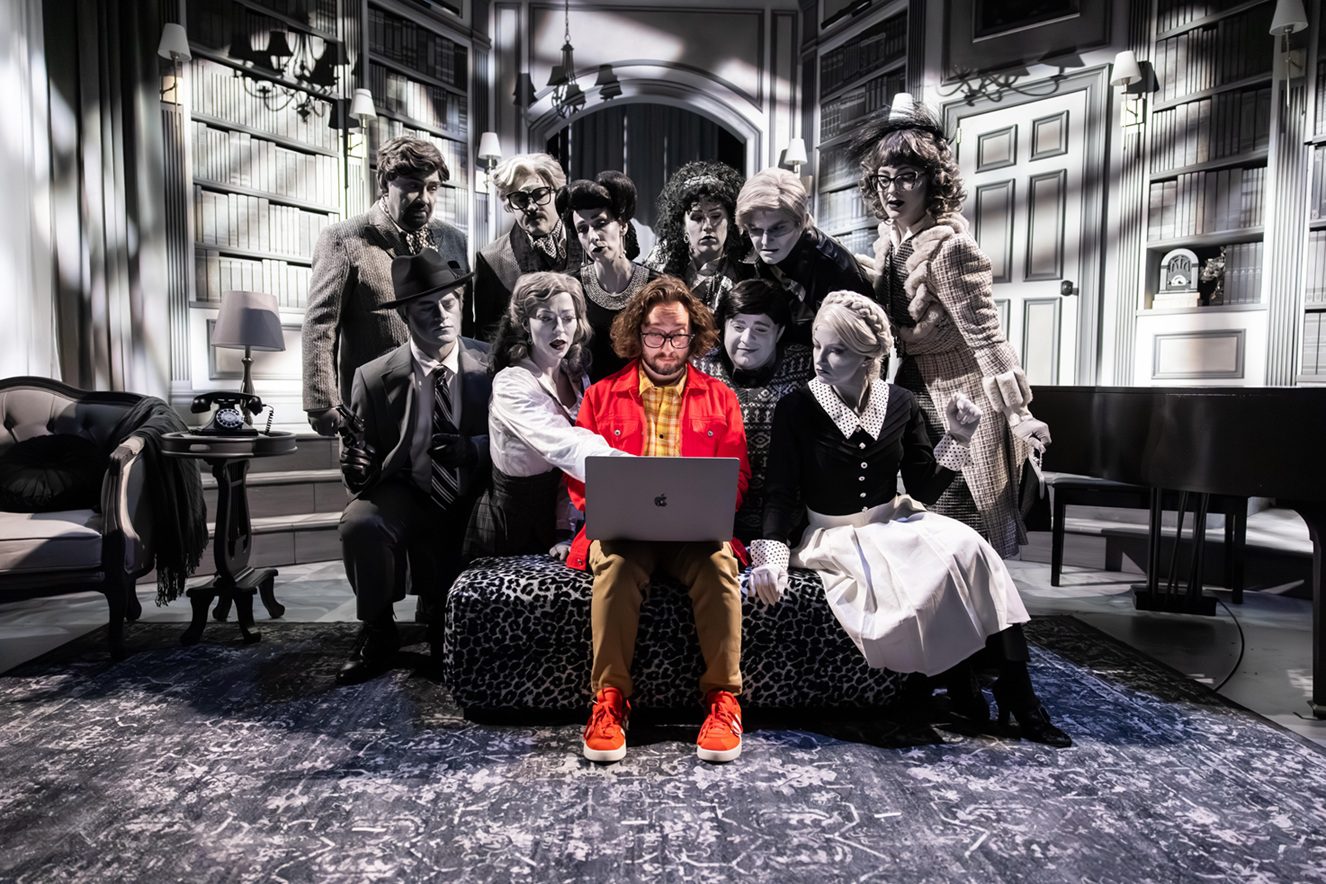The image is a theatrical scene featuring a group of people in a library setting. A man in a bright red jacket and khaki pants, wearing orange sneakers, sits in the center on a patterned bench with a laptop on his lap. Surrounding him are several individuals dressed in 1940s-style clothing, characterized by suits, hats, and dresses. The entire setting and the people around him are in grayscale, emphasizing the vivid color of the man's clothing. The backdrop includes bookshelves, a telephone, a lamp, and a grand piano. The characters' expressions suggest they are looking intently at the laptop screen.