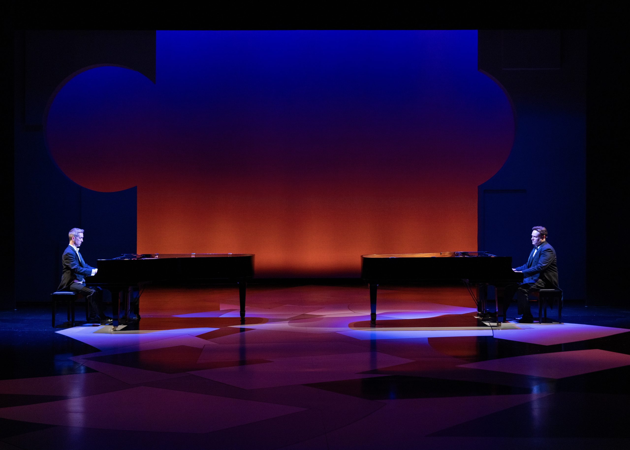 The image depicts a stage with two grand pianos positioned on opposite sides. Each piano has a person seated, facing each other. The stage is bathed in dramatic, colorful lighting with an orange backdrop that transitions to deep blue at the top. The floor features abstract geometric patterns illuminated with shades of blue and purple light. The individuals are dressed in formal black suits, each concentrating on their respective pianos. The overall atmosphere is serene and sophisticated, highlighting the concert-like setting.