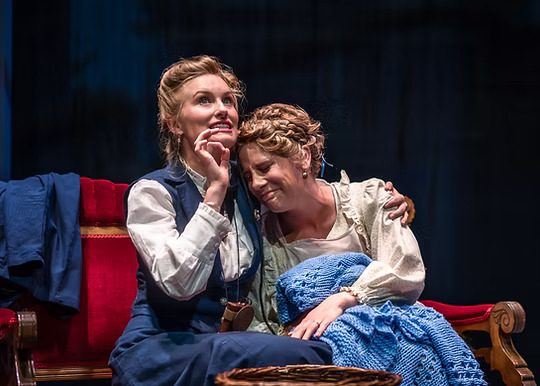 The image depicts a scene from the play "Silent Sky." Two women are seated on a red-upholstered wooden settee with intricately carved armrests, placed against a dark backdrop. The woman on the left is dressed in a blue dress with a white collar and appears to be gesturing upwards with an excited or pondering expression. Her light-colored hair is styled neatly. The woman on the right, wearing a white blouse with puffed sleeves, rests her head on the shoulder of the woman on the left, displaying a gentle expression. She is covered with a blue knitted blanket. A piece of blue fabric is draped over the armrest of the settee in the background.