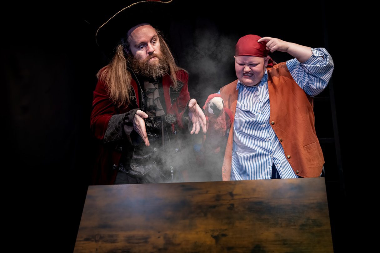 The image shows a theatrical scene featuring two actors in costume. On the left, an actor wears a dark, wide-brimmed hat and a deep red velvet coat, portraying a character with a beard and long hair. His hands are gesturing expressively in front of him, and he has a focused expression on his face. On the right, another actor wears a red bandana, a striped blue and white shirt, and a brown vest. This actor is making an animated facial expression, appearing to concentrate while pointing to their head. In between the actors, a small amount of smoky haze rises, adding a dramatic effect to the scene. The background is dark, highlighting the characters and their actions.