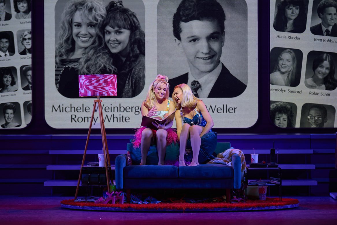 The image depicts a lively scene from the off-Broadway production of "Romy and Michele's High School Reunion." Six performers are on stage in a dynamic pose. The two central figures, wearing a shiny blue dress and a fluffy pink dress, stand at the front, holding hands raised triumphantly above their heads, smiling brightly. Surrounding them, four other performers strike playful poses; two on either side dressed in stylish costumes. The background consists of a large screen displaying colorful, geometric light patterns in shades of pink and blue. The stage is framed by curved arches, and the lighting enhances the vibrant and celebratory atmosphere.