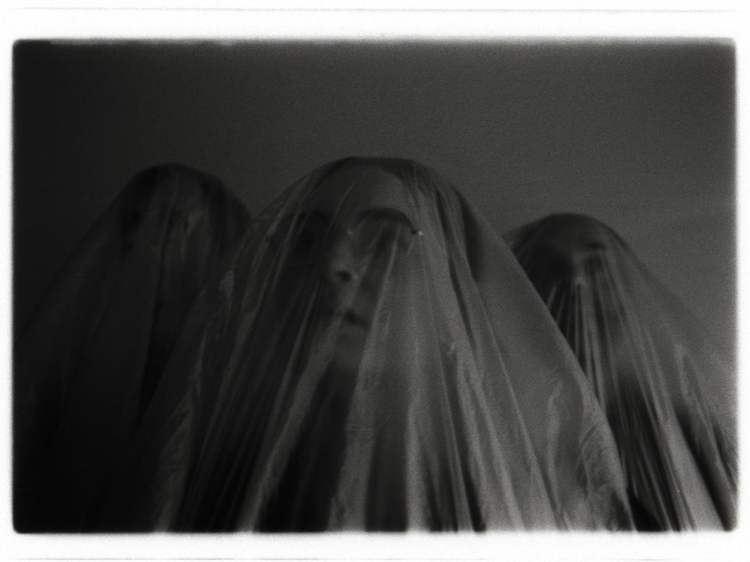 The black and white image shows three figures draped in transparent plastic sheets. They are centrally positioned, with the middle figure slightly more prominent. The plastic creates a flowing texture, obscuring the features of the figures beneath. The background is indistinct and softly blurred, enhancing the mysterious atmosphere. The image is framed with a subtle white border.
