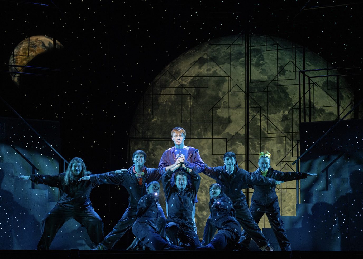The image depicts a theatrical scene with a group of performers on stage. The central figure, standing slightly elevated, is framed against a large backdrop depicting a celestial body, possibly a stylized moon, with geometric patterns overlaying it. The stage is set in a space-themed ambiance, with a starry background and soft, cool lighting that highlights the performers. Surrounding the central figure are seven individuals, dressed in dark outfits adorned with star motifs, suggesting an outer space or cosmic theme. They are posed dynamically, with outstretched arms creating a sense of movement and unity. The setting is framed by stage elements resembling stairs on both sides, continuing the starry pattern.