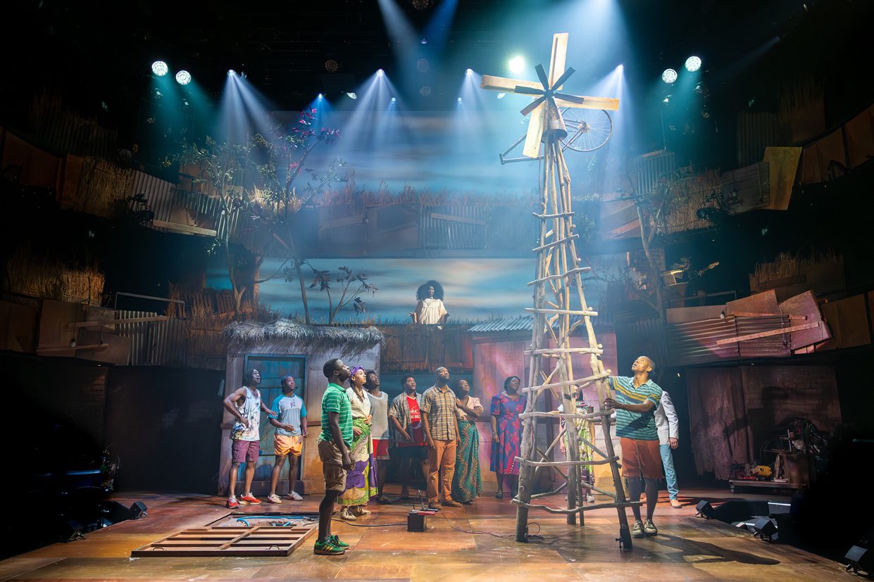 The image depicts a scene from a UK production of "The Boy Who Harnessed the Wind". The setting features a rustic stage design that conveys a rural environment. A tall, wooden windmill stands prominently in the center of the stage, constructed from crisscrossed wooden beams with a weathered, rustic look. Around the windmill, several performers are positioned, appearing to be engaged in an animated discussion or activity related to the windmill. The ensemble includes both male and female actors, some in colorful attire, contributing to a vibrant atmosphere. The background features a painted backdrop of blue skies with scattered clouds and silhouettes of trees, while structures resembling traditional African huts are partially visible. Soft, atmospheric lighting from overhead spotlights casts dramatic shadows and highlights the performers, enhancing the scene's emotional tone.